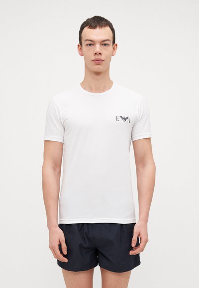 T-shirt blanc à manches courtes avec col rond et un petit logo noir sur la poitrine gauche. Matière en coton lisse contre un short sombre.