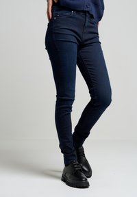 Tumši zili džinsu skinny bikses ar gludu tekstūru un pieguļošu dizainu, kas aprīkotas ar divām priekšējām kabatām un klasisku pogu aizdari.