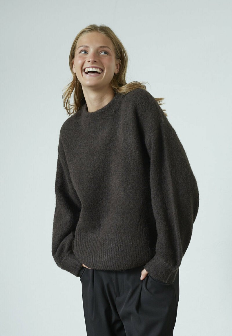 Brun sweater lavet af blødt, tekstureret stof med rund halsudskæring, oversize ærmer og ribbet kant. Bæres med sorte bukser.