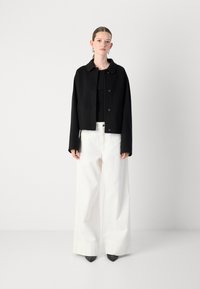 Zwarte kortvallende jas met een ronde kraag en knoopsluiting, gecombineerd met wijd uitlopende witte broek en taps toelopende zwarte hakken.