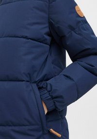 Tmavomodrá prešívaná bunda s elastickými manžetami a koženou nášivkou logo na rukáve. Hladký a jemný povrch.