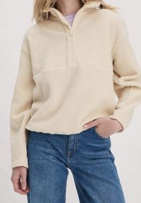 Sudadera de forro polar color crema con cuello de media cremallera, que presenta una textura suave y un panel en contraste en el pecho. Combinada con jeans azules.