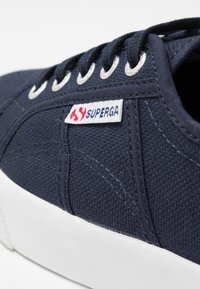 Tmavě modré plátěné tenisky s bílou podrážkou, s kovovými očky a bílou nálepkou s červeným a modrým brandingem "Superga".