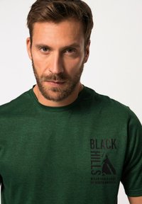 Zelené tričko s krátkým rukávem vyrobené z texturované tkaniny má malý černý grafický potisk na levém prsu, zobrazující hory a text.