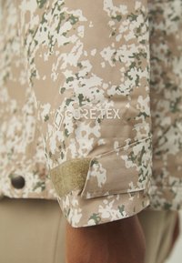 GORE-TEX-Jacke mit Ärmeln im hellbraunen Camouflage-Muster, ausgestattet mit einem strukturierten Verschluss und einem weichen, velcroähnlichen Streifen.