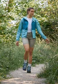 Himmelblau und türkisfarbene Windjacke mit Kapuze, weißes Shirt, beiges Shorts und dunkle Wanderschuhe, die einen Schotterweg in grünem Laub entlanggehen.