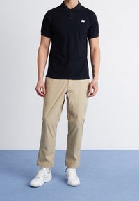 Camiseta polo negra con un pequeño logo blanco, combinada con pantalones chinos beige y zapatillas blancas. El conjunto tiene un aspecto casual y a medida.
