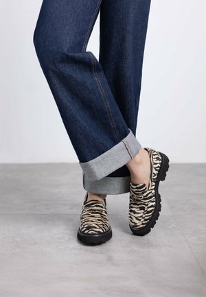 Zapatos slip-on de estampado animal en beige y negro con una parte superior texturizada, combinados con jeans de mezclilla enrollados con puños grises en una pose casual.