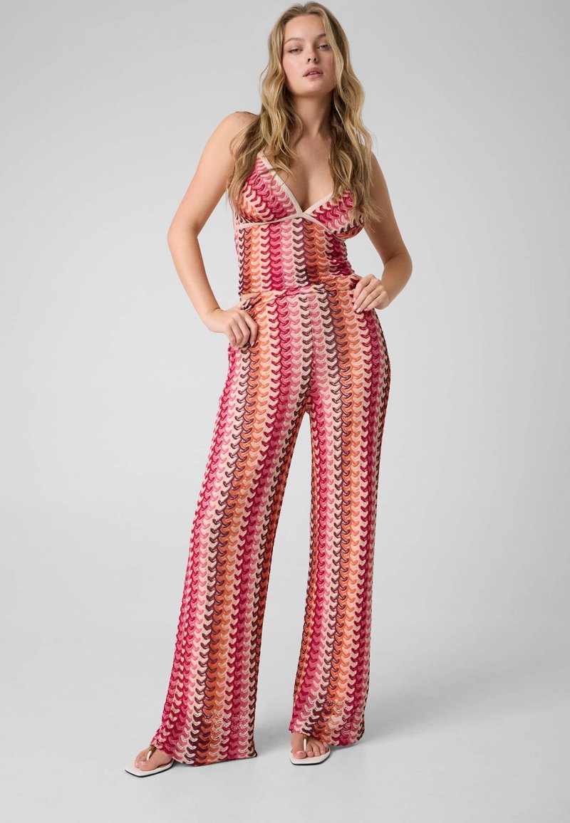 Femme portant une combinaison sans manches à col en V avec pantalon à jambes larges, présentant un motif rayé ondulé rose, rouge, orange et beige, debout les mains dans les poches.