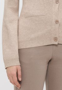 Bežs adīts kardigāns ar ribotām apakšmalām un aprocēm, ar mazu kabatu un plastmasas pogām priekšā. Pāris ar bežām biksēm.