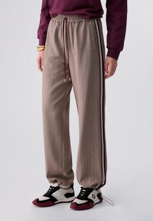 Pantalones beige de corte recto con rayas verticales y rayas laterales color granate, combinados con zapatillas negras y blancas y una camiseta de manga larga granate.