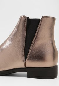 Metallinhohtoinen ruusukultainen nilkkurit, joissa joustavat sivupaneelit, matala korko ja sileä pinta. Varustettu pyöreällä varpaalla ja kestävä pohja.