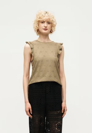 Femme aux cheveux blonds bouclés portant un haut beige sans manches à œillets avec des volants aux épaules et un pantalon en dentelle noire à motifs, debout sur un fond blanc.