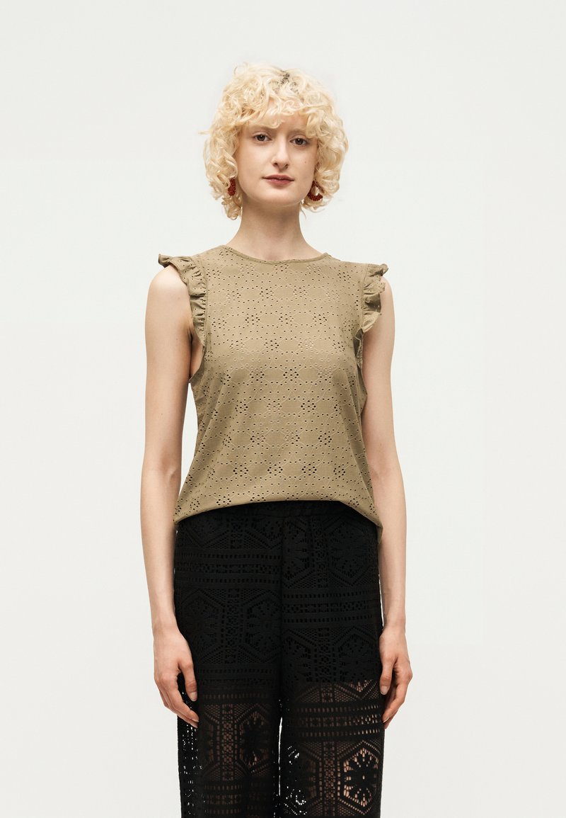 Γυναίκα με σγουρά ξανθά μαλλιά που φοράει ένα μπεζ αμάνικο τοπ με τρύπες και βολάν στους ώμους και μαύρο, δαντελένιο παντελόνι με σχέδια, στέκεται μπροστά σε λευκό φόντο.