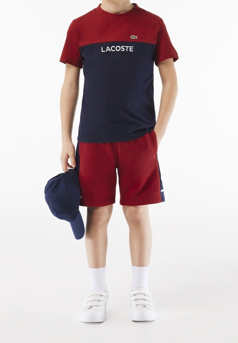 Enfant portant un t-shirt Lacoste rouge et marine et un short rouge, tenant une casquette marine, avec des chaussettes blanches et des baskets blanches.