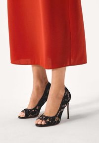 Jalat, joissa mustat pitsiset avoimet korkokengät ohuilla koroilla, yhdistettynä pitkä punainen hame, joka päättyy nilkkojen yläpuolelle.