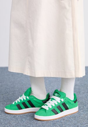 Zielone sneakersy Adidas Campus z czarnymi paskami, noszone z białymi prążkowanymi skarpetkami i beżową spódnicą na niebieskim dywanie.