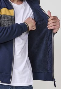Chaqueta azul marino con cremallera, panel texturizado en gris y acentos en amarillo. El interior forrado cuenta con un suave tejido de felpa. Puños y dobladillo acanalados.