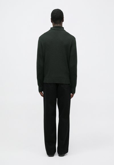 Σκούρο πράσινο πλεκτό πουλόβερ με ψηλό γιακά, συνδυασμένο με μαύρο παντελόνι χαλαρής εφαρμογής, από πίσω σε ανοιχτό φόντο.