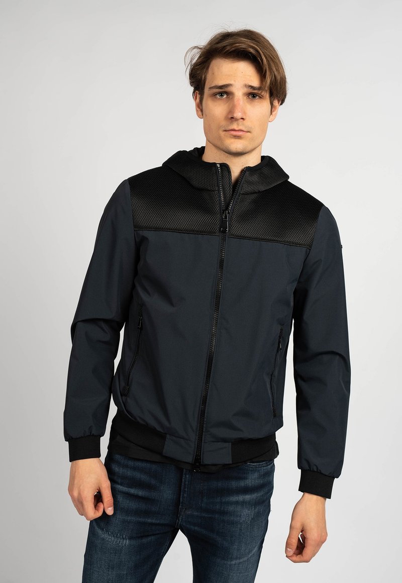 Giacca bomber nera e blu scuro con cappuccio, caratterizzata da dettagli in rete sulle spalle, polsini a coste e due tasche laterali.