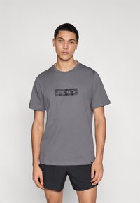 Γκρι βαμβακερό μπλουζάκι με στρογγυλή λαιμόκοψη, με ένα μαύρο ορθογώνιο λογότυπο στο στήθος. Φορεμένο με σκούρα σορτς. Ομαλή υφή.