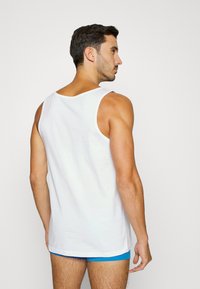 Débardeur en coton blanc avec un dos plongeant, un ourlet plat et de larges bretelles, porté sur un short bleu. La texture semble lisse.