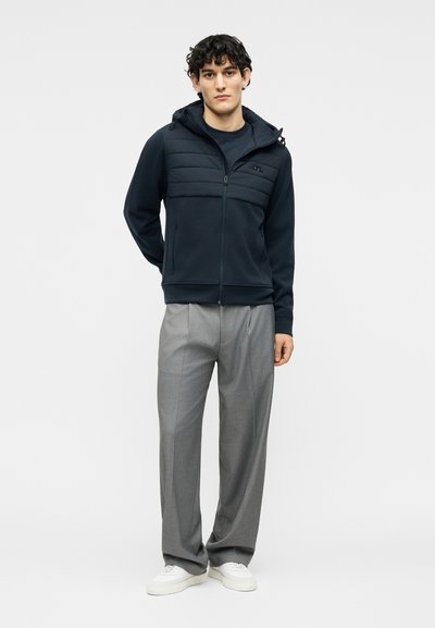 Νέος άνδρας που φοράει μαύρο καπιτονέ φούτερ με φερμουάρ, γκρι πλισέ παντελόνια και λευκά αθλητικά παπούτσια, στέκεται μπροστά σε απλό φωτεινό φόντο.