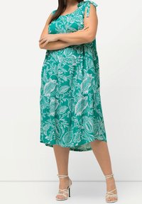 Kvinna som bär en ärmlös tealklänning med vita bladmönster och vita strappy högklackade sandaler, stående med armarna över kors.