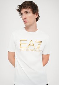 Hvit bomull t-skjorte med et stort gulllogo som leser "EA7 EMPORIO ARMANI", med klassisk rund hals og korte ermer.