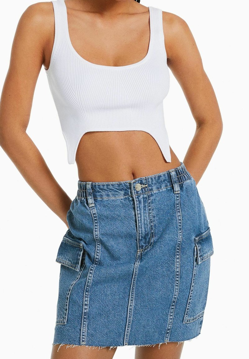 Bílé proužkované crop top s širokými ramínky, doplněný o světle modrou džínsovou sukni s náprsními kapsami a roztrhaným lemem. Oba kusy mají přiléhavý střih.
