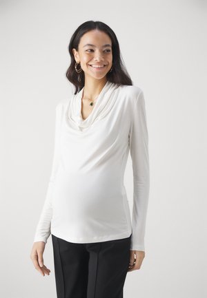 White long-sleeve top with a draped neckline, made of soft fabric, fitted through the torso, worn over black trousers.