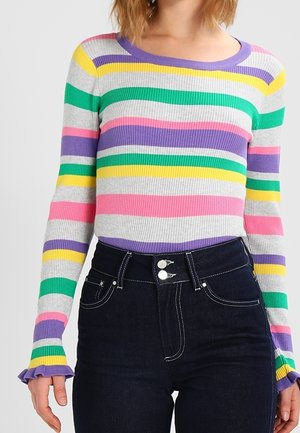 Frau trägt einen gerippten, mehrfarbig gestreiften Pullover mit Rüschen an den Ärmeln, kombiniert mit hoch taillierten, dunkelblauen Jeans mit doppelten Knöpfen.