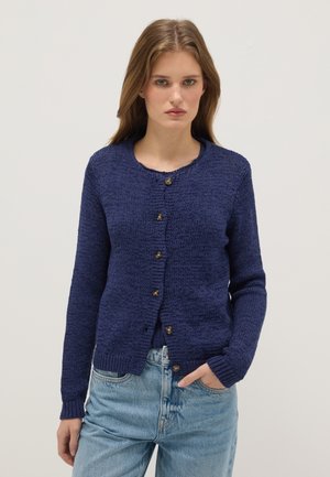 Junge Frau trägt eine marineblaue, geknöpfte Strickjacke mit langen Ärmeln und hellblaue Jeans, steht vor einem schlichten Hintergrund.