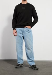 Uomo che indossa un felpa nera con testo bianco piccolo, jeans larghi blu chiaro e sneakers nere, in piedi con una mano in tasca.
