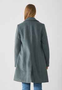 Cappotto in lana tessuta di un verde scuro con colletto a punte, vestibilità su misura e taglio medio, dotato di dettagli sulle cuciture e tasche laterali.