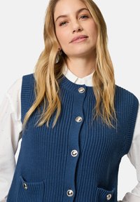 Blaues Strickwesten mit gerippter Textur, rundem Halsausschnitt und silbernen Knöpfen. Verfügt über zwei vordere Taschen. Trägt man über einem weißen Kragenhemd.