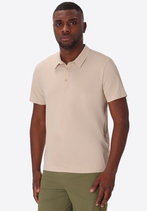 Uomo con capelli corti che indossa una polo beige a maniche corte e pantaloni verde oliva in piedi davanti a uno sfondo semplice.