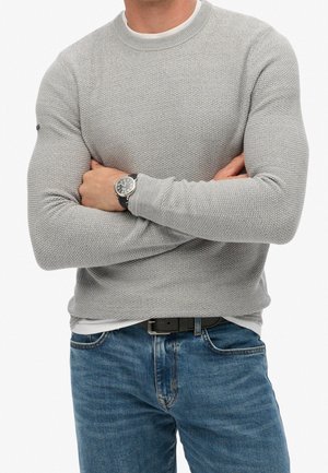 Mand iført en lysegrå struktureret sweater og blå jeans, med armene krydset, og med et sort ur på venstre håndled.