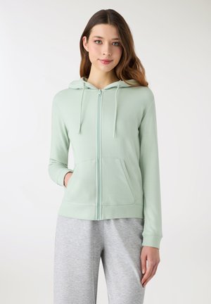 Felpa con cappuccio verde chiaro con zip, cappuccio con coulisse, tasche anteriori e polsini a coste. Indossata sopra pantaloni della tuta grigi, con una texture liscia.