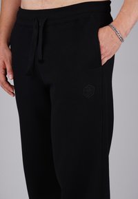 Close-up of person wearing black sweatpants with drawstring, hand in pocket, and silver chain bracelet on wrist against gray background.