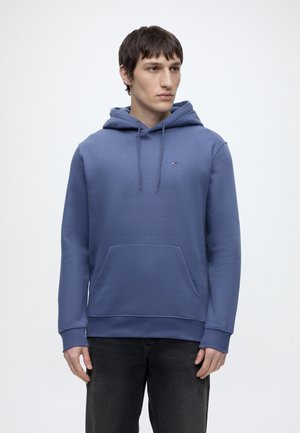 Junger Mann trägt einen blauen Hoodie mit Fronttasche und Kordeln, kombiniert mit schwarzen Jeans, stehend vor einem schlichten weißen Hintergrund.
