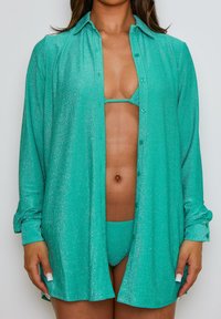 Camicia turchese scintillante con maniche lunghe e bottoni, indossata sopra un bikini coordinato, con un design minimalista e una texture liscia.