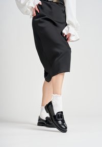 Scarpe in pelle nera con una larga fibbia, indossate con calze alla caviglia bianche a volant. L'outfit presenta una gonna nera e una blusa bianca strutturata.