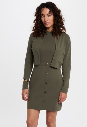Vestido verde oliva con un top acanalado ajustado, sobrecortado con una chaqueta corta y detalles de botones en la parte delantera. Cuenta con mangas largas y dos bolsillos en el pecho.
