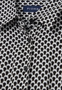 Black and white patterned button-up shirt featuring a collar, fabric with a repetitive design, and black buttons. Brand label visible.
