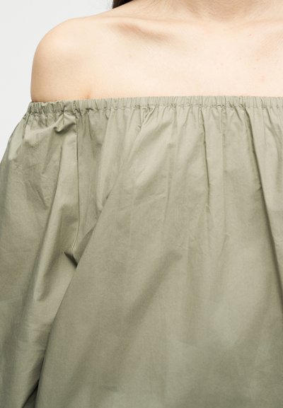 Γυναίκα που φοράει φαρδιά, χακί πράσινη μπλούζα έξω από τον ώμο με ελαστικό ντεκολτέ, που δείχνει τις κλείδες και την περιοχή του άνω στήθους.