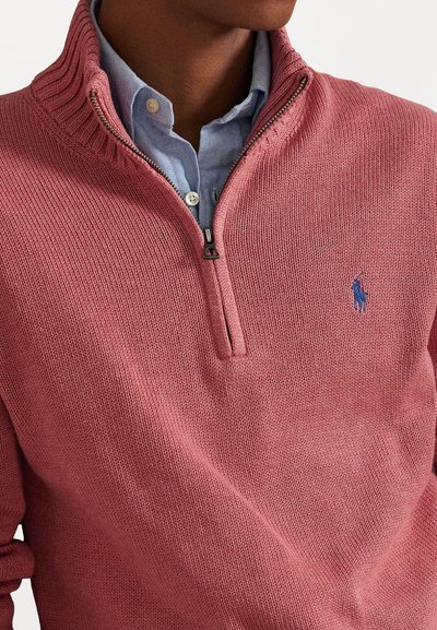 Κόκκινο πλεκτό πουλόβερ με κλείσιμο φερμουάρ στον γιακά και ριγωτή υφή, τοποθετημένο πάνω από ένα ανοιχτό μπλε πουκάμισο με κουμπιά; διαθέτει μπλε λογότυπο στο στήθος.