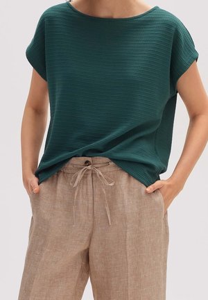 Vrouw draagt een donkergroene, geribbelde top met korte mouwen, ingestopt in beige linnen broek met trekkoord, handen in de zakken.