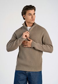 Beiger gerippter Strickpullover mit halben Reißverschluss-Kragen; lange Ärmel; lockere Passform; kombiniert mit blauen Jeans; weiche Textur.
