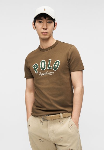 Καφέ βαμβακερό μπλουζάκι με λευκό και πράσινο κείμενο "POLO Ralph Lauren", συνδυασμένο με ανοιχτό μπεζ παντελόνι διακοσμημένο με κεντημένα σχέδια σκύλων.